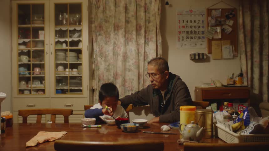 A father sits at a table and laughs and helps his young son as he eats