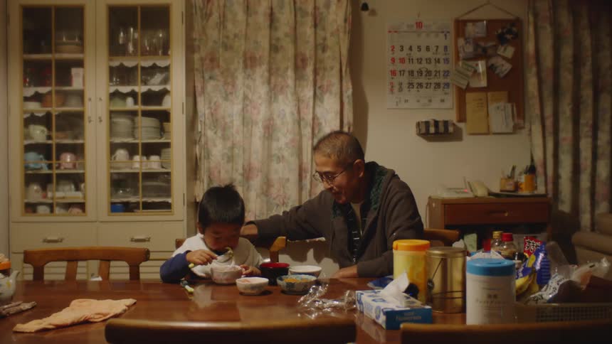 A father watching his young son eat food from a bowl while sitting at a dining table