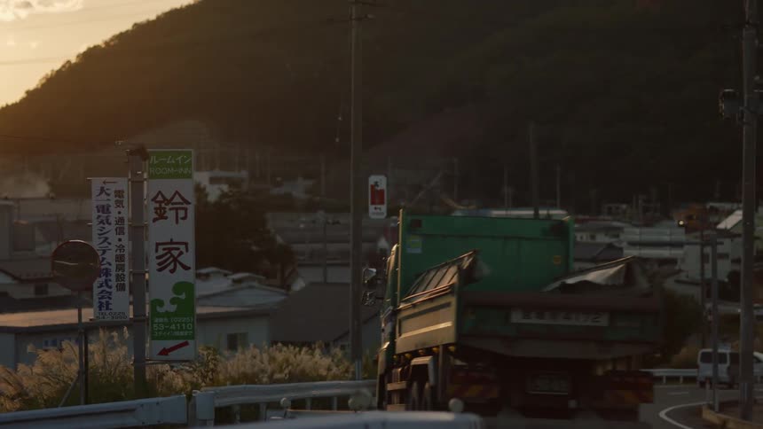 Vehicles travel along a road through a residential neighborhood at the base of a forested mountain in the evening