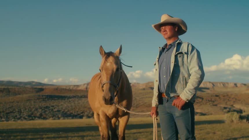 Cowboy stands with horse near house with arid scrubland and mountains in the background
