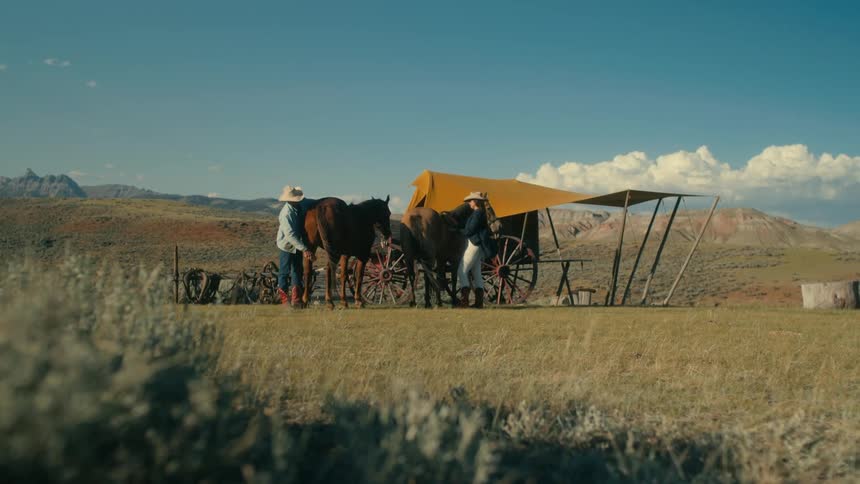 A cowboy brushing a horse next to a parked covered wagon