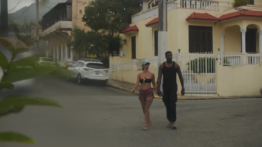 A man and a woman are holding hands as they cross a street and walk away from a building with cars parked in front