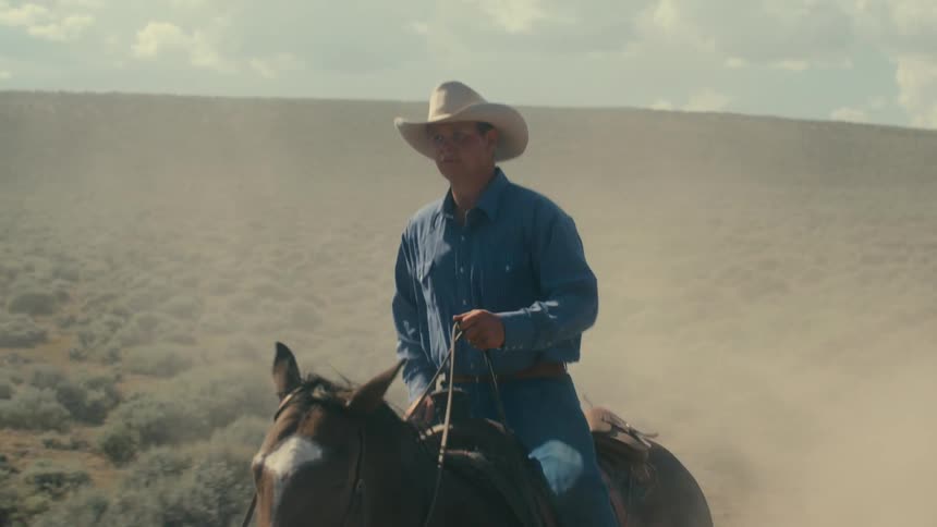 Man with brown hat blue shirt and jeans rides brown horse in the field