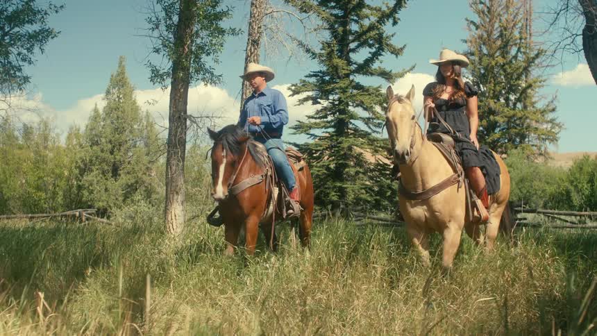 A man and woman ride horses through a field surrounded by a wooden fence during the day