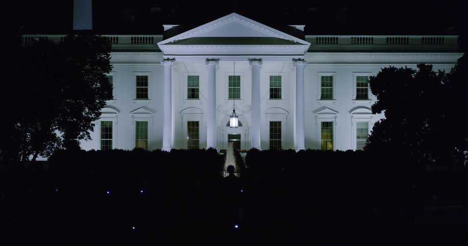 Silhouettes of crowd move around near hedge enclosing the white house at night