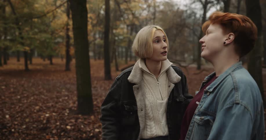 One woman greets another with a kiss and they walk through woods in park together