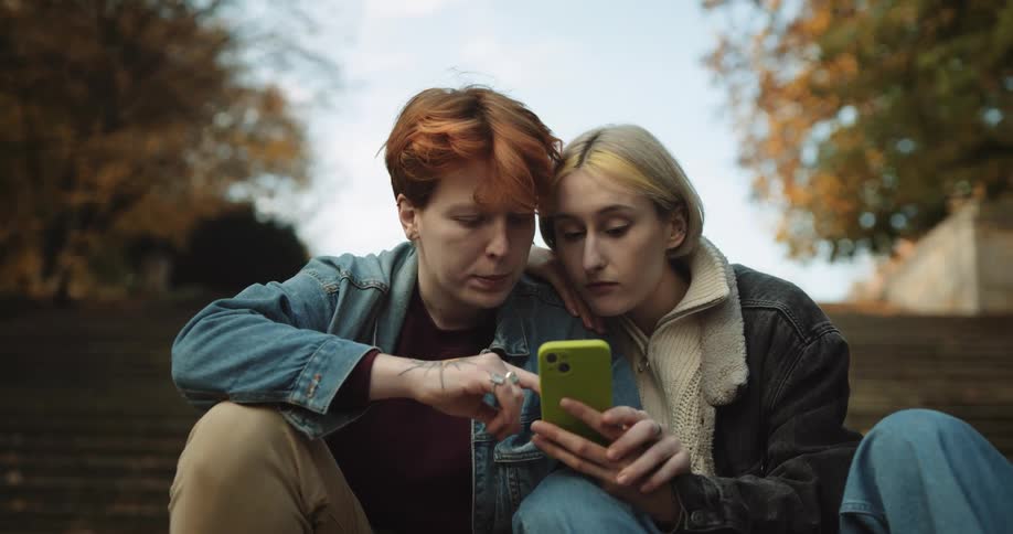 Lesbian couple wearing denim jackets jeans and pants sits on stairs using and look at smartphone