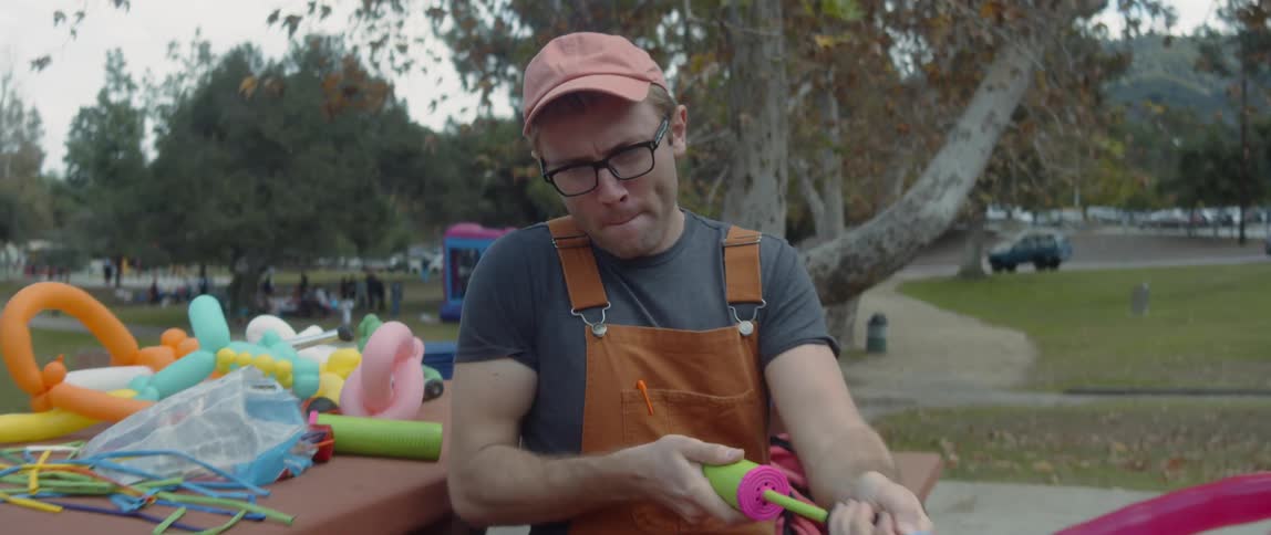 A man sits by a crowded park and blows up balloons