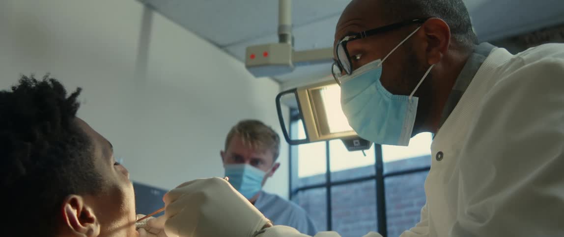 A dentist positions a light and uses a dental mirror to examine the mouth of a patient