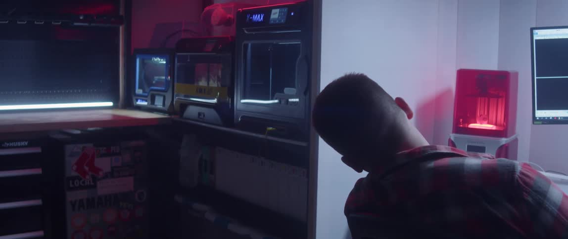 A man is sitting at a desk with a laptop computer and a large curved monitor as he moves his head to look at a machine in the next room