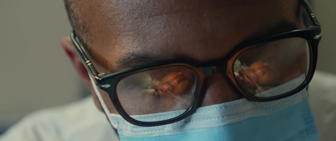 A man wearing glasses and a surgical mask is looking down as he nods his head