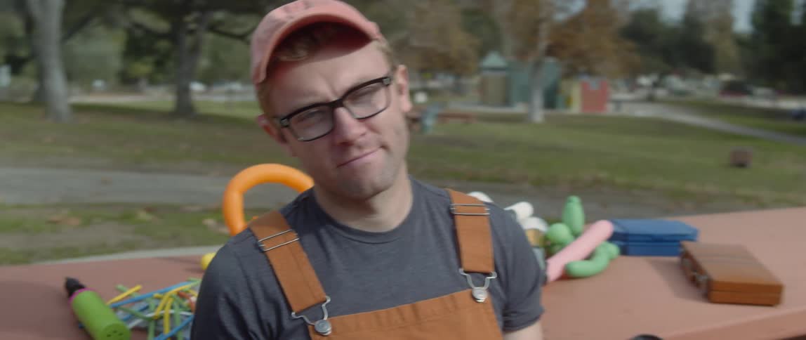 Man wearing overalls sits on a park bench filled with party supplies