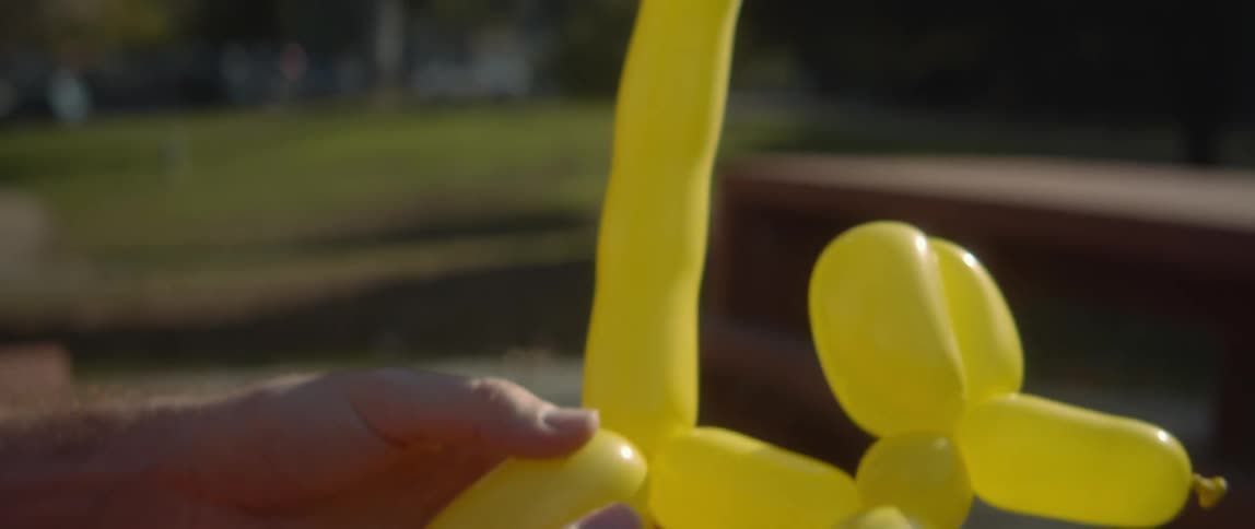 The sun shines on a man outside twisting a yellow balloon into an animal with his hands