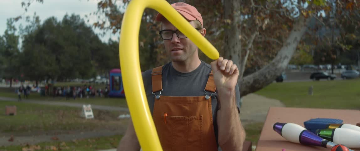 A man sits at a park with a party going on and blows up a balloon and gives it to someone