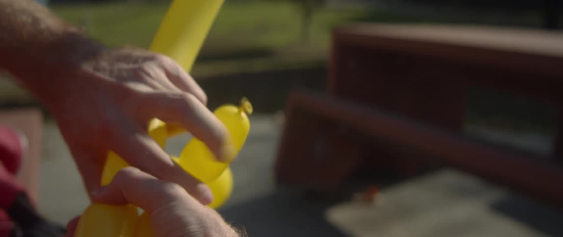 The sun shines on a man outside twisting a yellow balloon into an animal with his hands