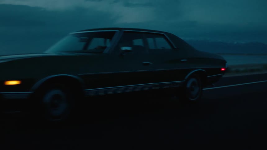 An automobile with headlights on travels on a road along the coast at night and there are mountains in the distance