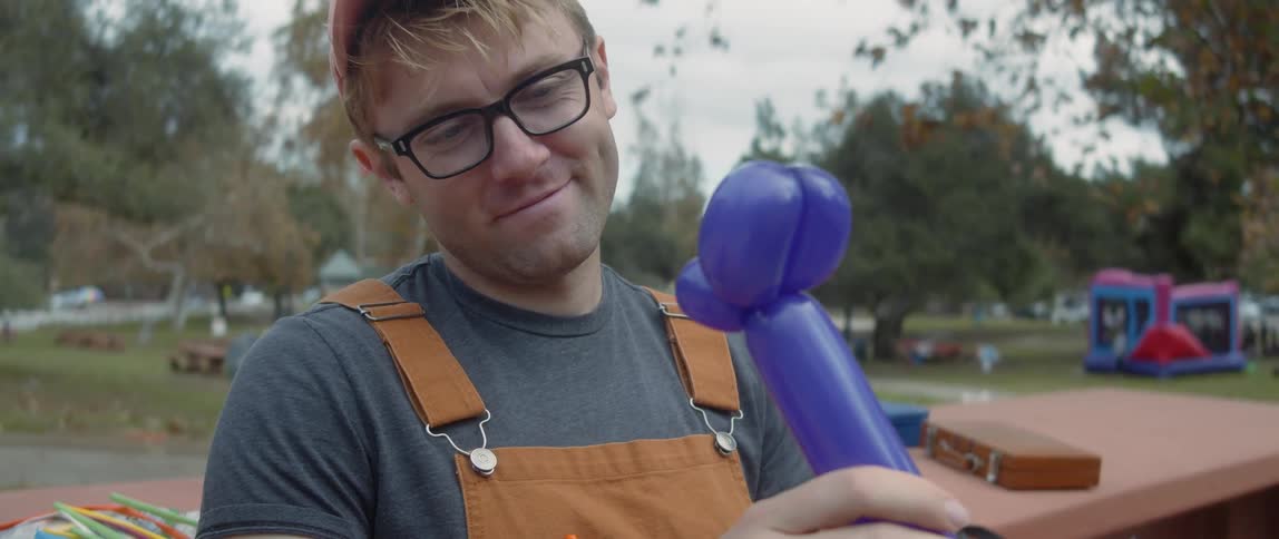 A young white man is blowing up balloons at a public park