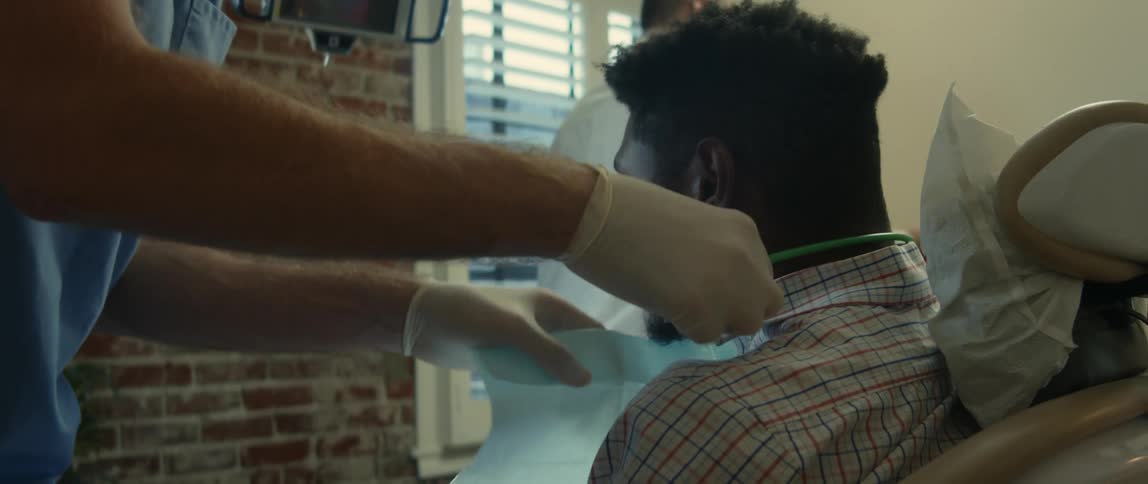 A patients sits in a dentists chair as a man puts a bib around his neck and the dentist washes his hands