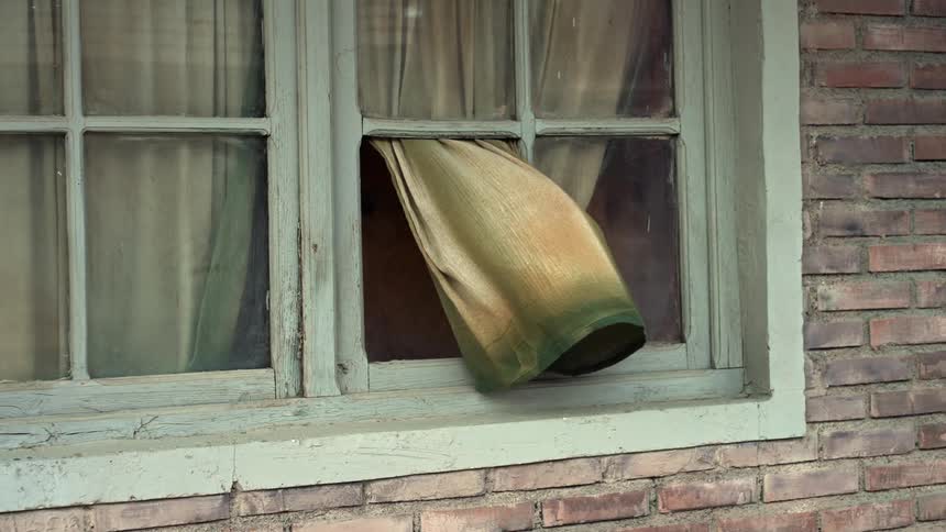 A discolored curtain in an old broken window is blowing in the wind