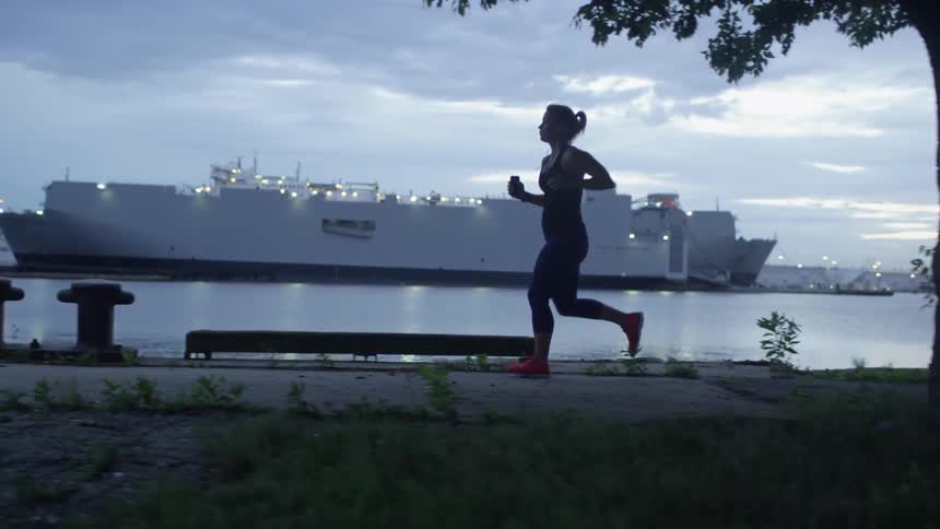 A woman is jogging near the water with a large ship