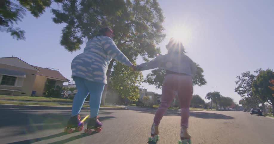 Two teenage girls hold hands and roller skate down a neighborhood street