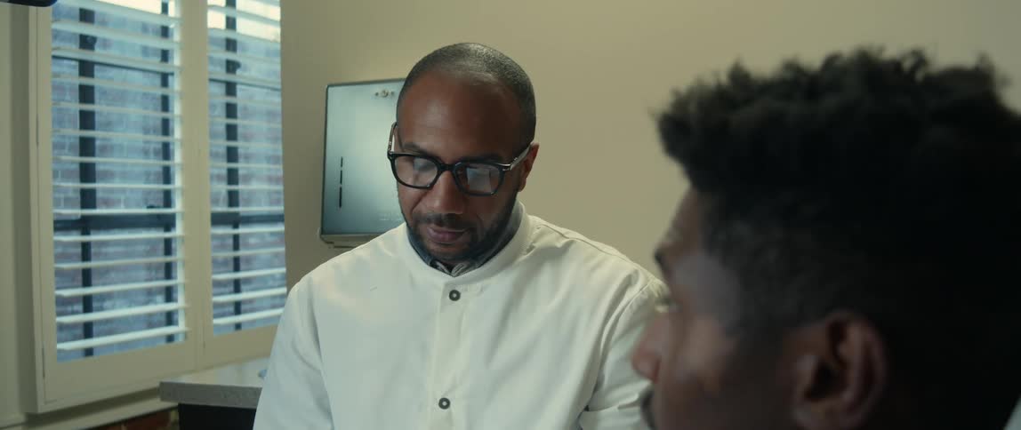 A man wearing a white shirt is sitting in an office looking at a notebook and talking to a younger man