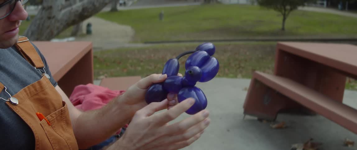 A man is sitting outside twisting a blue balloon into an animal with his hands