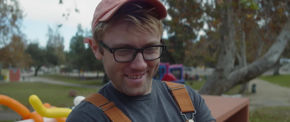 A man smiling while making a purple balloon animal in an outdoor park