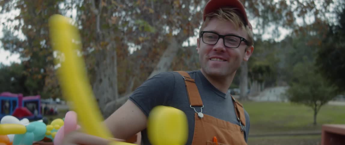 A young male balloon artist hands a yellow balloon dog to someone and waves at them