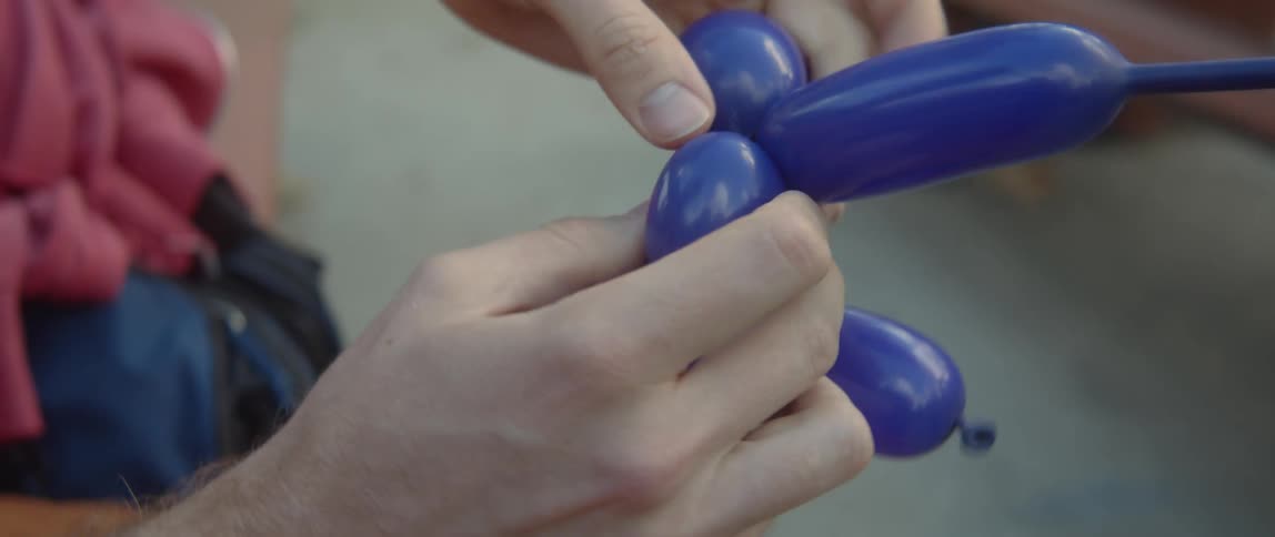A man is sitting outside twisting a blue balloon into an animal with his hands