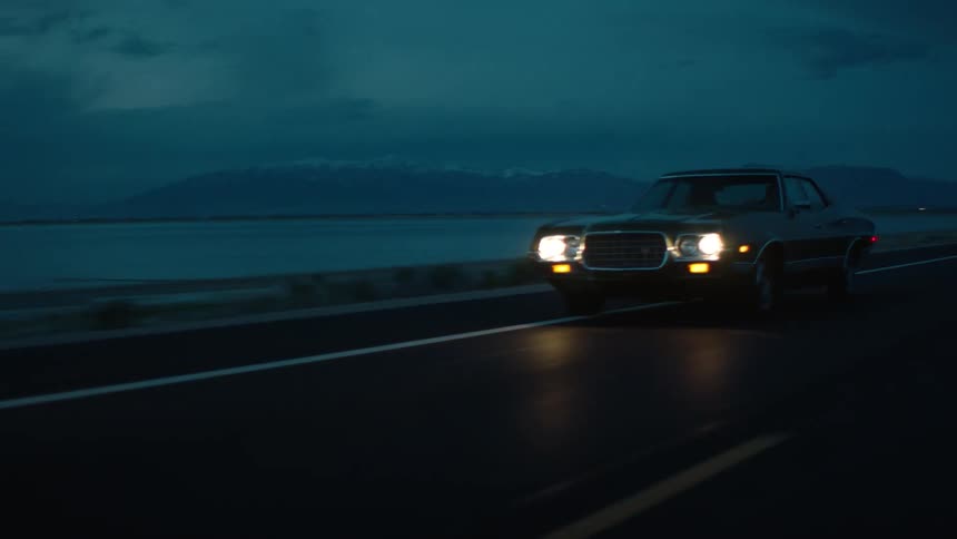 Car drives along road paralleling water on foggy night with mountains on the horizon