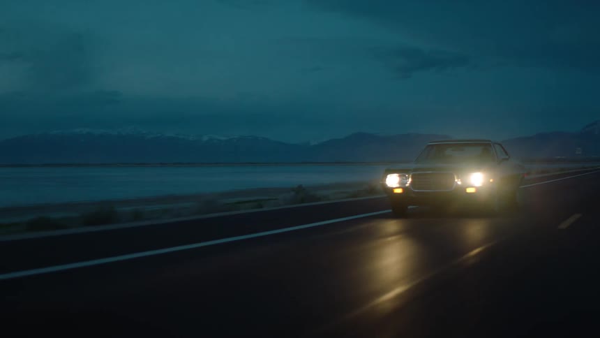 Person drives car along road near shore with snow covered mountain in the background