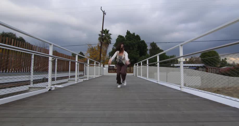 A teenage girl runs across a walk way and turns around and swings her arms in the air and throws them out to her side