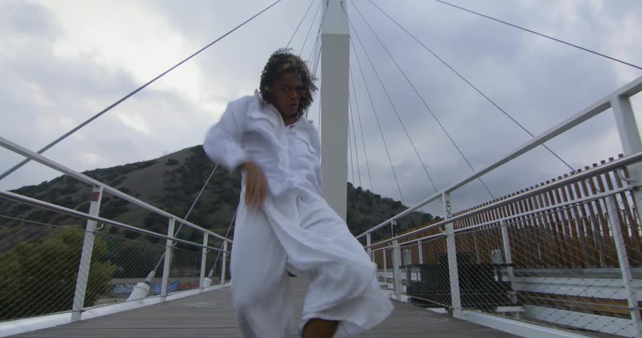 Young performer wearing white dances fluidly walking forward over a suspension bridge