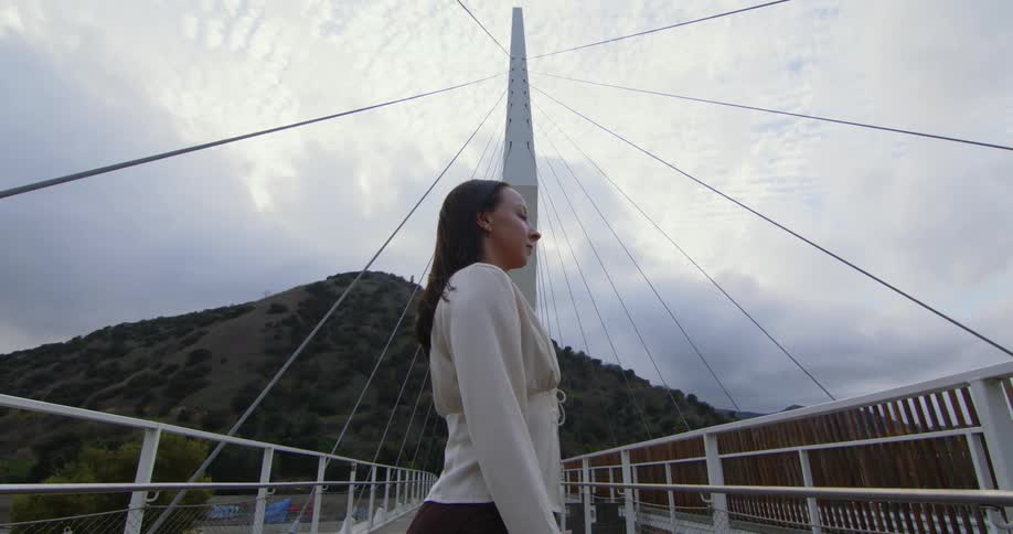 Woman stands between rails on cable foot bridge under an overcast sky near hill