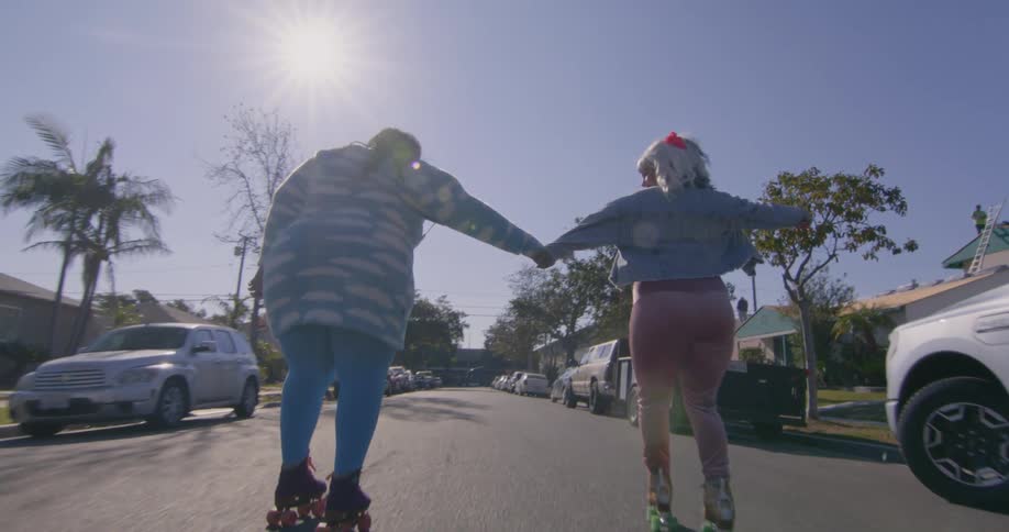 Two women hold hands while roller skating down street between parked vehicles on sunny day