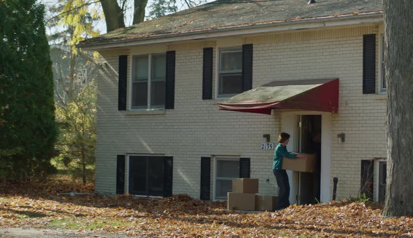A woman holds boxes while standing in the doorway of a house before another person retrieves one of the boxes and carries it inside