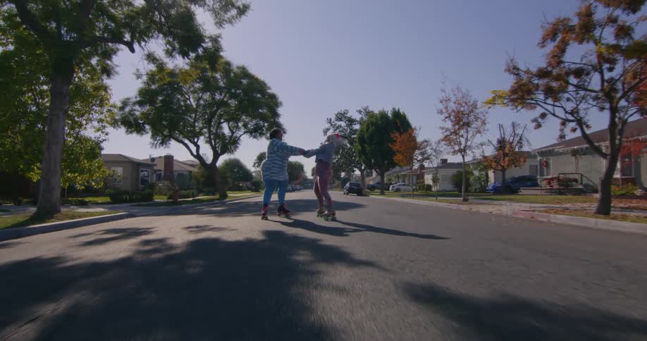 Two teenage girls are on roller skates in the middle of a neighborhood street holding hands and spinning in a circle