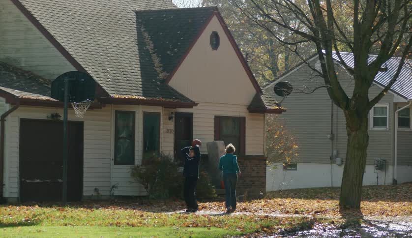 A teenage boy is outside throwing a basketball through the hoop as a woman runs to the house
