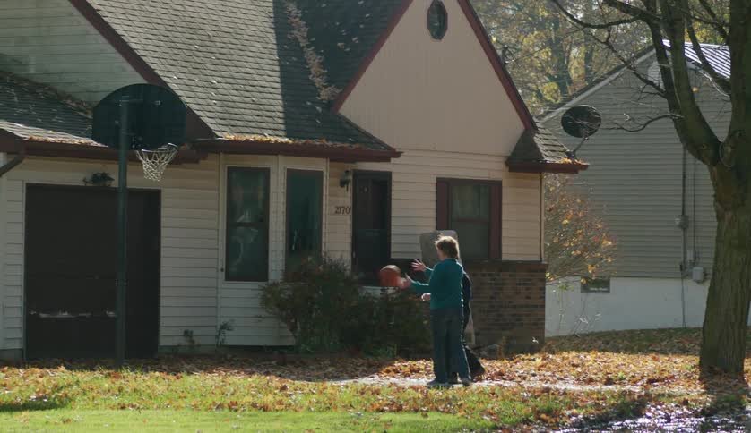 A teenage boy is standing outside with his mother as he shoots baskets and she takes a shot and misses