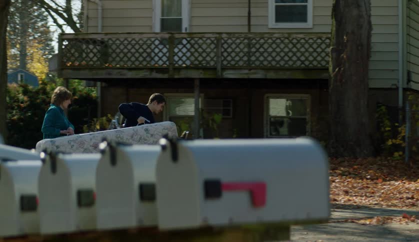 Woman and man carry mattress across street and fallen leaf strewn yard before stopping to rest on porch of house