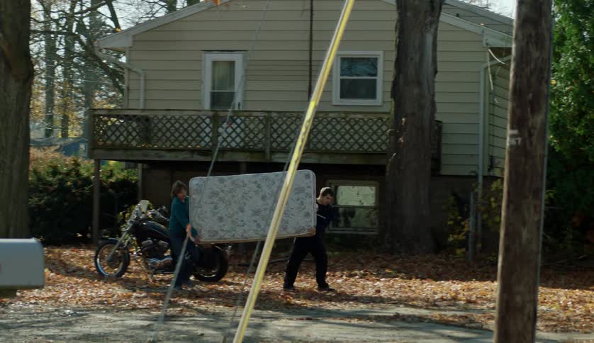 Two young men are carrying a twin mattress across a residential neighborhood into another house in autumn