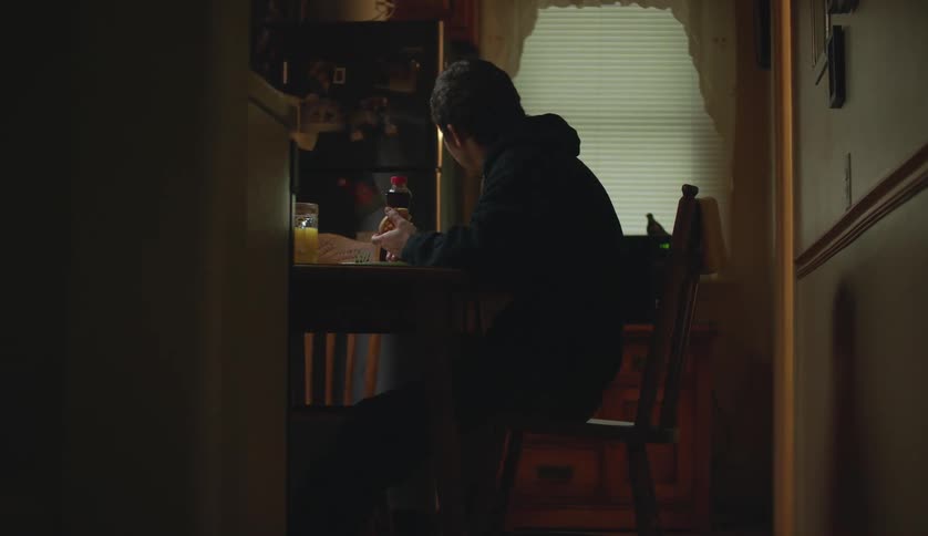 Man wearing black hoodie and pants sits at table in the kitchen talking to woman