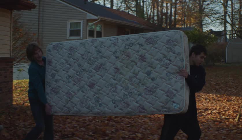 A woman and a young man carry a mattress out of a house on an autumn day