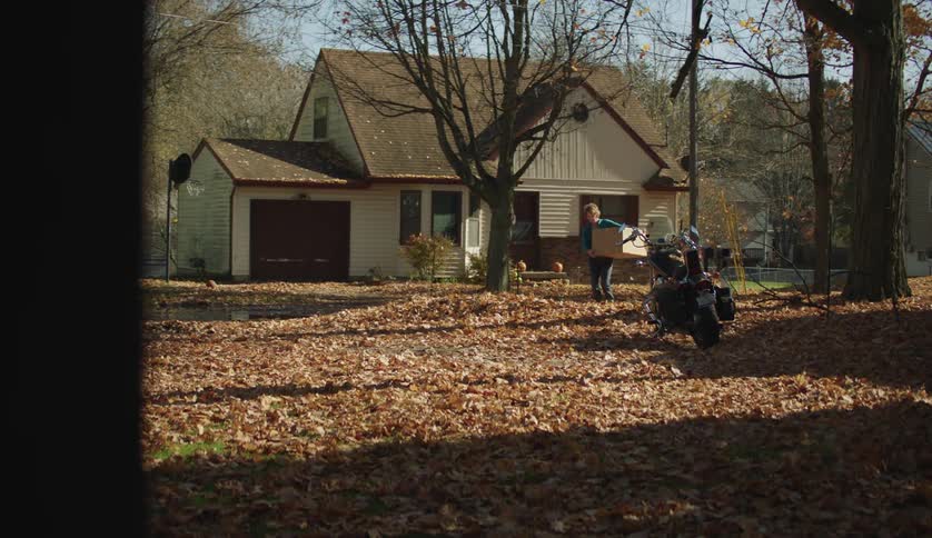 Middle aged woman in blue jacket and jeans carries a package out of house in the countryside