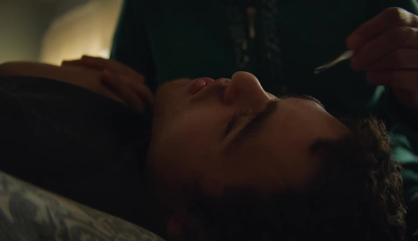 A teenage boy is lying on his bed as his mother tweezes his eyebrows