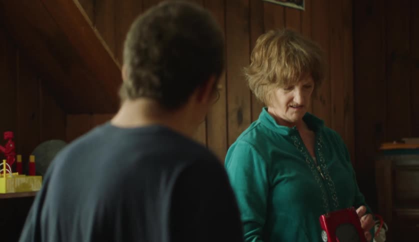 A mother looks at a toy car in her teenager sons room and he holds his arms out to his side in confusion