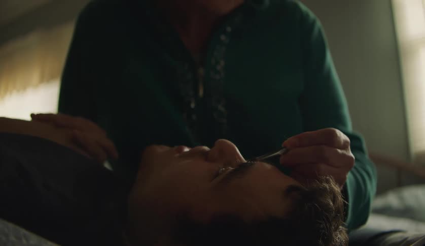 Young man lies on his back and talks to older woman as she tweezes his eyebrows
