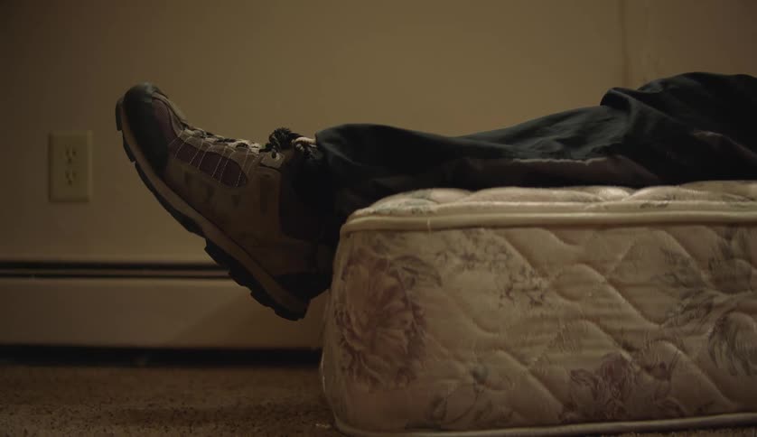 Person lies on bare mattress on the floor wearing sneakers