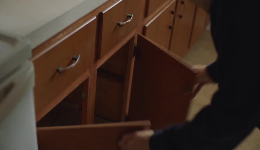 A young man searches through the empty cabinets and drawers of a home kitchen during the day